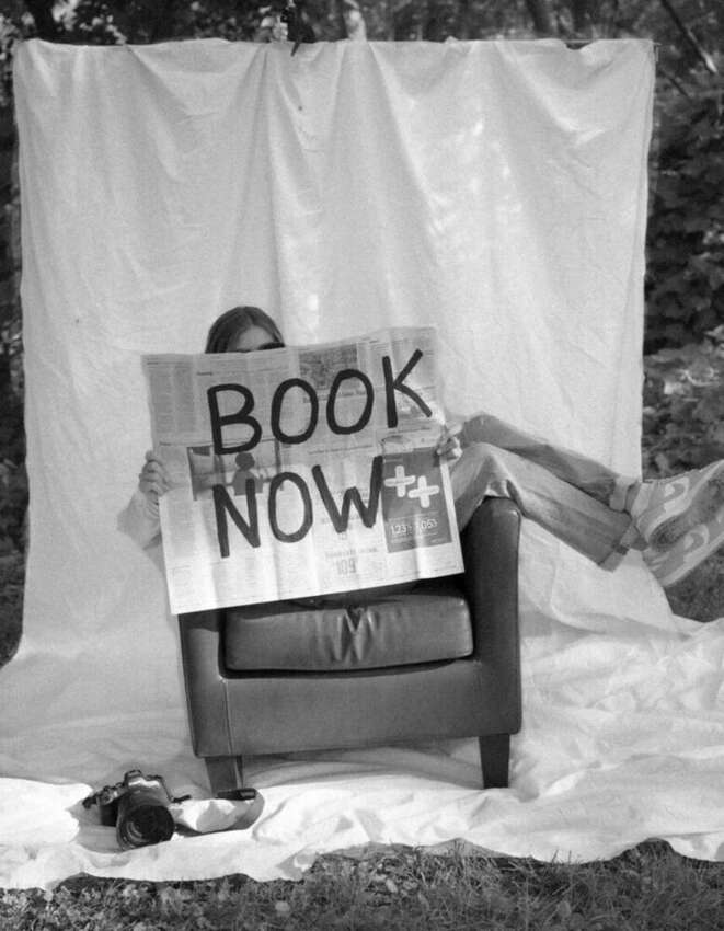 Black and white photo of Isabella holding a newspaper sign that reads Book Now