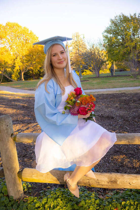 Senior girl in a graduation cap and gown sitting on a fence holding flowers