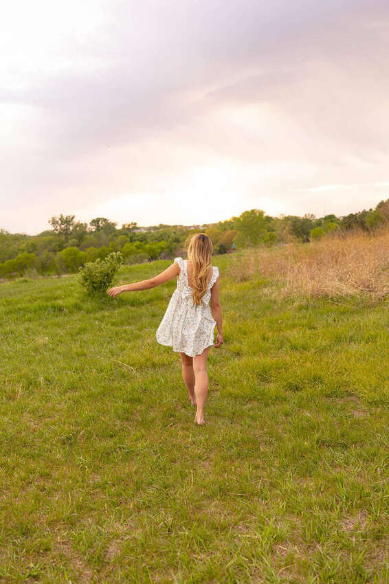 Senior girl in a white dress walking through a field, back view, pink sunset sky